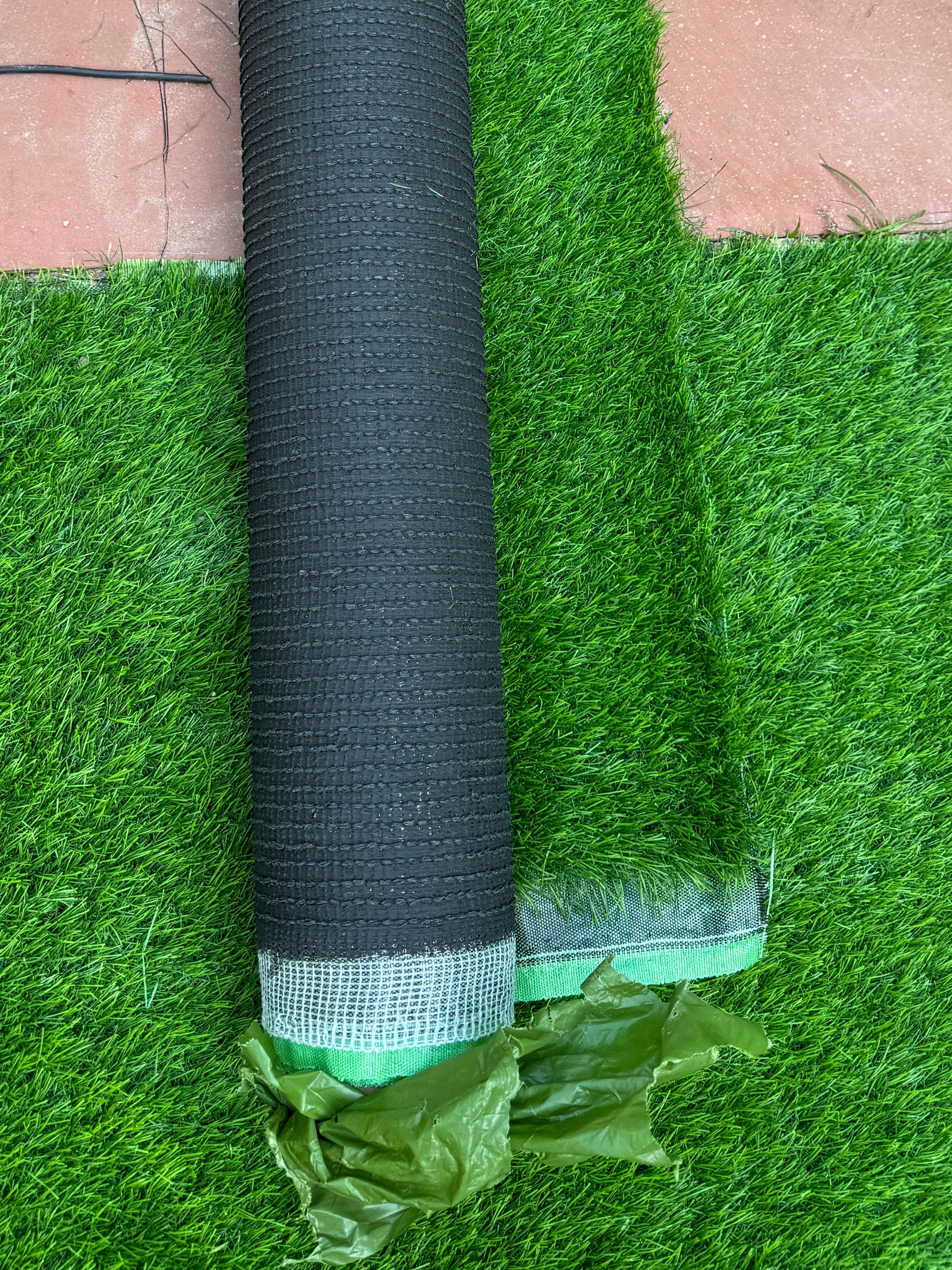 Full frame image of artificial, green grass lawn being laid by block paved patio with red and black square tiles, rolls of fake turf, elevated view, focus on foreground